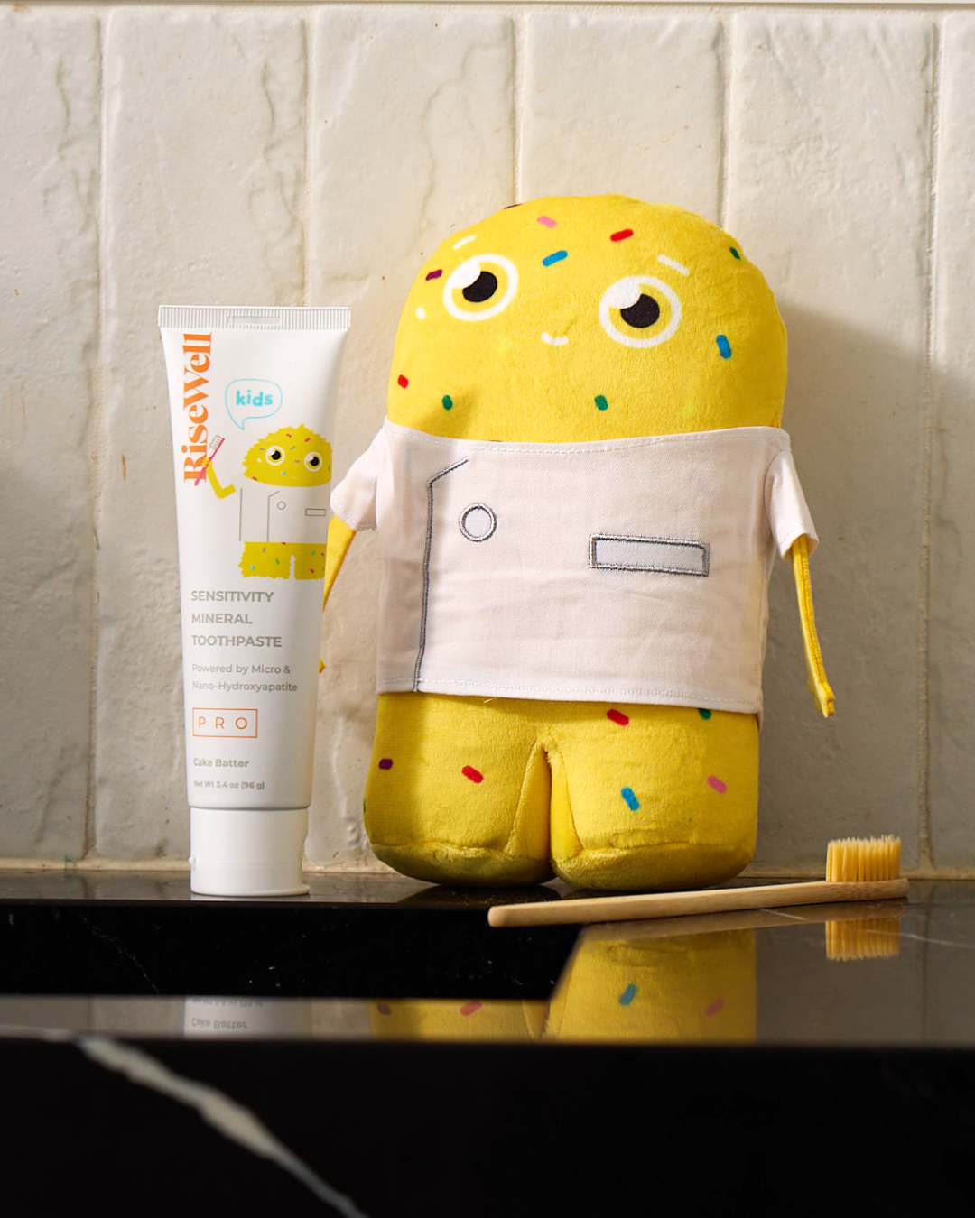 Yellow plush toy with a white shirt and colorful sprinkles next to a tube of RiseWell toothpaste on a reflective surface.
